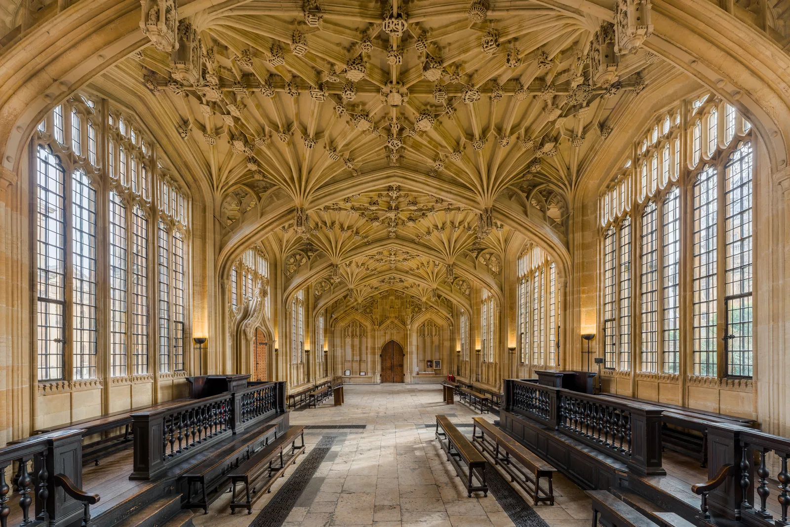 The Divinity School in Oxford, looking east along the hall beneath its carved fan vault, oak benches down either side of a flagstone floor.